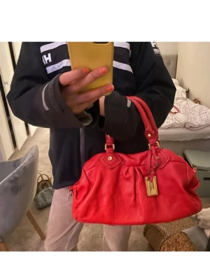 As cool Röd Marc by Marc Jacobs handväska - Cool röd handväska från Marc by Marc Jacobs i mjukt läder med guldiga metalldetaljer och dubbla handtag. Väskan har en rymlig form och dekorativ tag framtill. Perfekt för dig som vill sticka ut med en färgstark accessoar. Är precis imsmord med läderbalsam