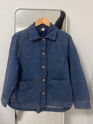 Mörkblå jeansjacka - Stilren mörkblå jeansjacka med klassisk krage, knäppning framtill med kopparfärgade knappar och tre stora fickor. Rak passform och tillverkad i denim. Perfekt för en avslappnad och trendig look.