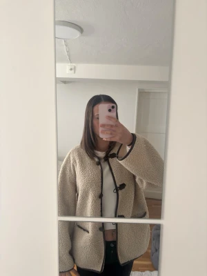 Beige teddyjacka med svarta detaljer - Mysig beige teddyjacka med svarta kantband och knäppning framtill. Jackan har en oversized passform och två fickor framtill. Perfekt för kalla dagar och ger en trendig look med sin fluffiga struktur.