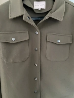 Olivgrön overshirt från KIDS ONLY - Stilren olivgrön overshirt från KIDS ONLY med klassisk krage, tryckknappar och två bröstfickor.