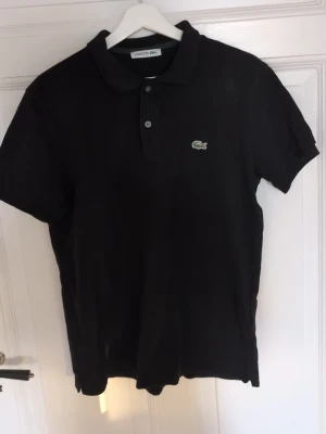 Svart polotopp från Lacoste - Klassisk svart polotopp från Lacoste med den ikoniska krokodilloggan på bröstet. T-shirten har korta ärmar och knappar vid halsen. Perfekt för dig som gillar stilrena och sportiga plagg.