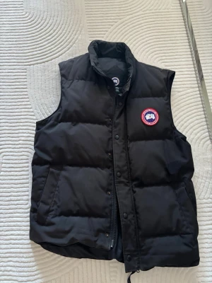 Svart dunväst från Canada Goose - Snygg svart dunväst från Canada Goose med klassisk logotyp på bröstet. Västen har hög krage, dragkedja och knappar framtill samt två fickor på sidorna. Perfekt för lager-på-lager och håller dig varm under kyliga dagar. Äkta såklart