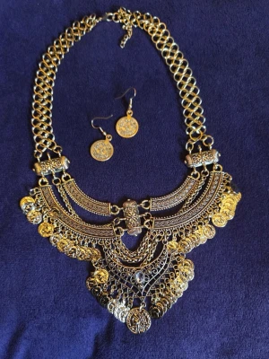 Guldigt halband och örhängen  med mynt vintage indisk bohem goth alternative - Säljer ett maffigt nytt och oanvänt guldigt halsband med matchande örhängen. Halsbandet har flera lager med detaljerade mönster och hängande mynt, vilket ger en bohemisk och unik look. Perfekt för att lyfta vilken outfit som helst. Materialet är metal - zinklegering i guldfärg.