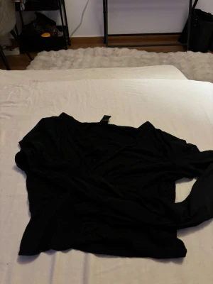 Svart omlottopp med lång genomskinlig ärm - Snygg svart omlottopp med lång genomskinlig ärm och v-ringad hals. Toppen har en enkel och stilren design som passar till många olika looks. Perfekt för dig som gillar minimalistisk stil.