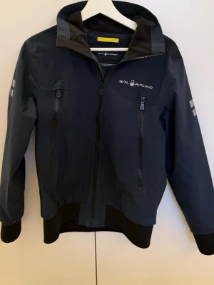 Svart vindjacka från Sail Racing - Stilren svart vindjacka från Sail Racing med huva, hel dragkedja och två stora fickor framtill. Jackan har logga på bröstet och reflexdetaljer på ärmarna. Materialet är vattenavvisande och jackan har en sportig, modern passform.