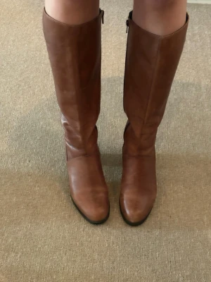 Bruna knähöga läderstövlar - Snygga bruna stövlar i äkta läder med knähögt skaft och dragkedja på sidan. Klassisk design med rundad tå och låg klack, perfekta till jeans eller kjol. Stövlarna har en stilren look och passar till många olika outfits. Från Vagabond. Storlek 37! Säljer för 300kr