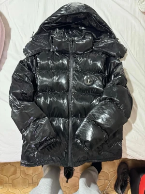 Svart pufferjacka från Trapstar - Säljer en svart glansig pufferjacka från Trapstar med stor huva och broderad logga på ryggen. Jackan har en patch med Trapstar-märke på bröstet, dragkedja framtill och elastiska muddar vid ärmarna. Perfekt för kalla vinterdagar och riktigt street.