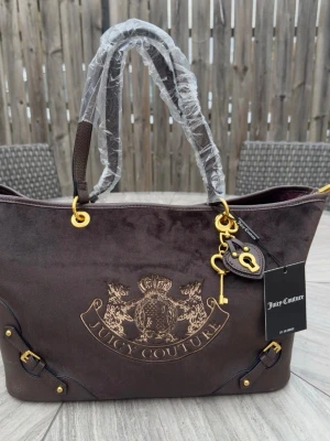 Juicy Couture brun sammetsväska - Mörkbrun handväska i sammet från Juicy Couture med broderad logga framtill, guldfärgade metalldetaljer och dekorativt hänglås med nyckel. Väskan har dubbla handtag och snygga läderdetaljer i hörnen. Perfekt för dig som gillar lyxig och trendig stil.