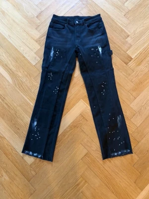 Svarta raka jeans med färgstänk - Coola svarta jeans med raka ben och unika vita färgstänk och slitningar. Jeansen har klassisk femficksdesign och hällor för bälte. Perfekta för dig som vill sticka ut med en edgy streetstil.