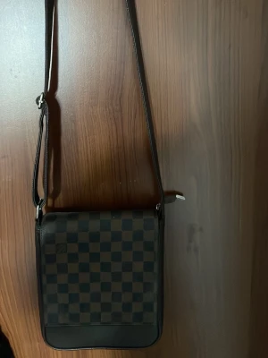 Svart axelväska med rutmönster - Snygg svart axelväska med justerbar rem och klassiskt rutmönster på framsidan. Väskan har en fyrkantig form och dragkedjeficka på baksidan. Perfekt för dig som vill ha en stilren och praktisk väska i vardagen.