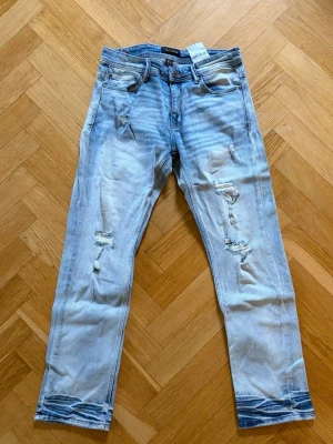 Ljusblå slitna jeans - Säljer ett par ljusblå jeans med slitningar och hål på benen för en trendig look. Jeansen har fem fickor, normal midja och smal passform. Perfekta för dig som gillar en avslappnad och modern stil.