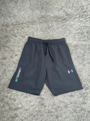 Gråa shorts från Under Armour (reflex) - Snygga gråa shorts från Under Armour med färgglada loggor och textdetaljer (reflex). Shortsen har elastisk midja med snörning och två sidofickor. Perfekta för träning eller chill. Materialet känns som mjuk polyester. Hela settet finns i min profil som en egen annons.