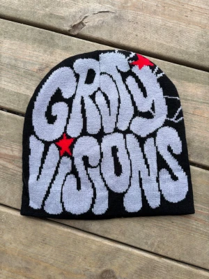 Svart mössa GRVTY VISIONS tryck - Säljer en svart mössa med stort grått GRVTY VISIONS-tryck och röda stjärnor. Mössan har en avslappnad passform och är stickad i mjukt material, perfekt för dig som vill sticka ut med streetwear-vibe.
