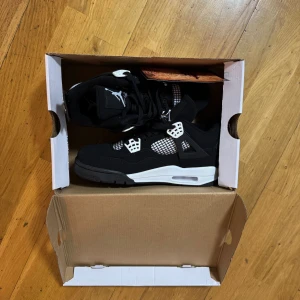 Svarta Air Jordan 4 Retro sneakers - Säljer ett par svarta Air Jordan 4 Retro sneakers med vita detaljer och meshpaneler. Skorna har klassisk snörning, ikonisk Jumpman-logga på hälen och en chunky sula. Materialet är en mix av syntet och mesh för skön komfort och stilren look.