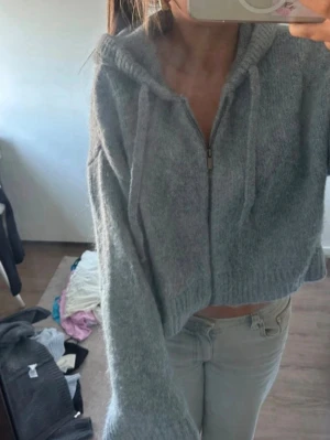 Zara stickad tröja  - Mysig croppad hoodie i ljusgrå färg med dragkedja framtill och huva med snören. Tillverkad i ett fluffigt, stickat material som känns riktigt skönt. Perfekt för en chill och avslappnad stil. Passar till jeans eller mjukisbyxor för en casual vibe.