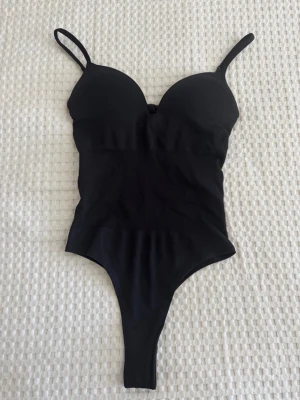 Svart body med tunna axelband - Snygg svart body med tunna justerbara axelband och vadderade kupor. Bodyn har en smickrande passform och stringmodell baktill. Tillverkad i stretchigt material som formar sig efter kroppen. Perfekt att styla under skjorta eller kavaj. Aldrig använt🥰