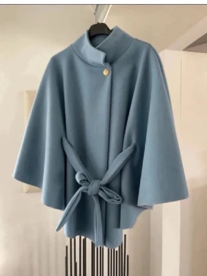 Ljusblå cape-kappa med knytband ny - Stilren ljusblå cape-kappa med knytband i midjan och en dekorativ guldknapp vid halsen. Kappan har vid passform, hög krage och vida ärmar för en modern look. Perfekt för dig som vill ha en trendig och elegant ytterjacka i mjukt tyg.