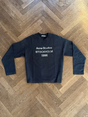Acne Studios Long Sleeve - Cool svart sweatshirt från Acne Studios med vit text 'Acne Studios STOCKHOLM 1996' på bröstet. Klassisk rund halsringning och långa ärmar. Perfekt för en avslappnad och trendig look. 175 på bild, passar någon runt 175-180