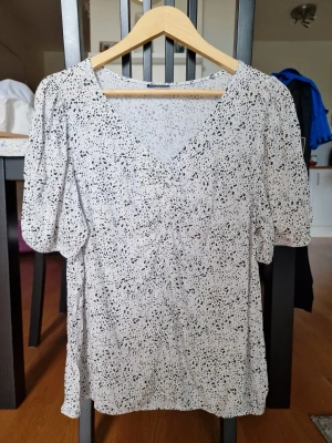 Vit blus med svart prickmönster  - Trendig vit blus från Beloved (Pepco) med svart prickigt mönster. Blusen har v-ringning och puffiga trekvartsärmar som ger en cool och avslappnad look. Tillverkad i ett lätt och luftigt material, perfekt för att styla med jeans eller kjol. Från viskos.