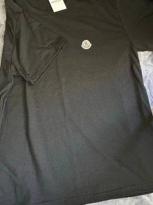 Svart Moncler t-shirt med logga - Snygg svart t-shirt från Moncler med klassisk rund hals och Moncler-logga på bröstet. Tillverkad i mjuk bomull för en bekväm känsla. Perfekt till jeans eller shorts för en clean och stilren look. Strl L/XL
