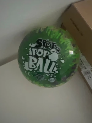 Grön Splatter Pop Ball - Säljer en grön Splatter Pop Ball från Mondo. Bollen är genomskinlig med gröna och lila färgstänk och har en cool pop-effekt när man kastar den. Perfekt för lek både inne och ute. Texten 'Splatter Pop Ball' syns tydligt på framsidan.