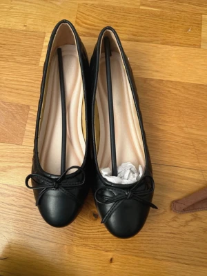 Svarta quiltade ballerinaskor med rosett - Stilrena svarta ballerinaskor med quiltade sidor och en söt rosett framtill. Skorna har rund tå och låg klack. Utsidan är i konstläder och insidan är ljusbeige. Perfekta för en trendig och klassisk look.nya