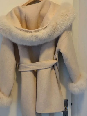 Beige kappa med tillhörande vantar - Stilren beige kappa med stor huva och fluffiga pälsdetaljer på huva och ärmslut. Kappan har knytskärp i midjan och två fickor framtill. Perfekt för dig som vill ha en mysig och trendig look under kalla dagar.