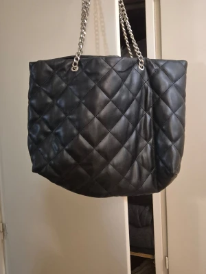 Svart quiltad shoppingväska med kedja - Stilren svart shoppingväska i quiltat mönster med silverfärgade kedjehandtag. Väskan har en mjuk, vadderad känsla och en rymlig form som rymmer mycket. Perfekt för dig som gillar en klassisk men trendig look.