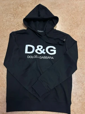 Svart D&G hoodie med logga - Snygg svart hoodie från Dolce & Gabbana med stor vit D&G-logga på bröstet. Klassisk huva och ribbade muddar vid ärmslut och nederkant. Aldrig använd, skriv i chatten om du vill se bild hur den sitter på och vikt och längd