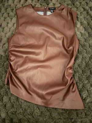 Brun ärmlös topp i fuskskinn MISU - Säljer en brun ärmlös topp från new yorker i mjukt fuskskinn. Toppen har rund halsringning, dragkedja bak och asymmetrisk nederkant. Snyggt figurnära passform och trendig look som sticker ut.