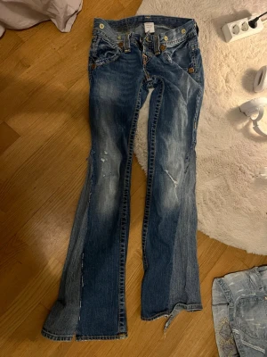 Blå slitna bootcut jeans från True Religion - Ett par blåa jeans från True Religion som jag har sytt om till bootcut. (Dem var tidigare straight leg) Det finns slitningar och slitna detaljer på benen. Jeansen har klassiska kontrastsömmar, fem fickor och knappar med logga. Materialet är denim i bomull och passformen är utställd nertill. Säljer denna eftersom att den inte passar mig längre. 