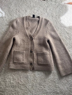 Beige stickad kofta med fickor - Mysig beige stickad kofta med v-ringning och tre knappar framtill. Koftan har två stora fickor fram och ribbade muddar vid ärmslut och nederkant. Från lindex