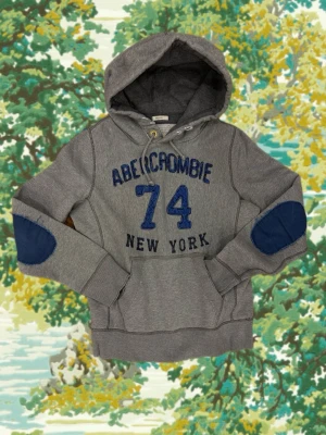  Abercrombie & Fitch hoodie - Snygg grå hoodie från Abercrombie & Fitch med blåa detaljer, broderad text och siffran 74 på bröstet. Hoodien har en stor huva, känguruficka och blåa armbågslappar. Tillverkad i mjukt bomullsmaterial, perfekt för chill dagar.