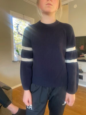 Mörkblå stickad tröja med vita ränder - Mörkblå stickad tröja med två vita ränder på varje ärm. Tröjan har rund halsringning och långa ärmar. 