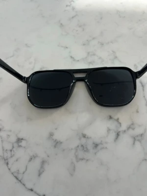 Svarta solglasögon med retroform - Snygga svarta solglasögon med klassisk retroinspirerad form och mörka glas. Bågarna är i plast och har en stilren look med en liten silverdetalj vid näsryggen. Perfekta för att höja din stil på soliga dagar.