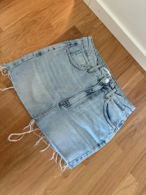 Ljusblå jeanskjol med fransig kant - Trendig ljusblå jeanskjol i klassisk femficksmodell med rå, fransig kant nedtill. Kjolen har bälteshällor, knapp- och dragkedjestängning samt slitna detaljer för en avslappnad look. Perfekt för dig som gillar en casual och cool stil.