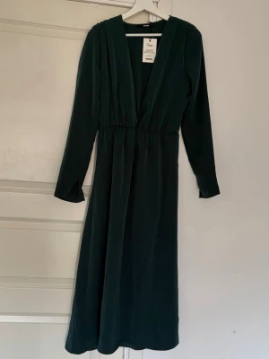 Mörkgrön långklänning från BikBok - Snygg mörkgrön långklänning från BikBok med djup v-ringning och långa ärmar. Tillverkad i 100% återvunnen polyester. Perfekt för dig som gillar stilrena och eleganta plagg med en modern touch.