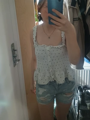 Vit prickig linnetopp med volang - Supersöt vit linnetopp med svarta prickar och volangkant nertill. Smockad överdel och breda axelband med volangdetalj. Toppen har knappar framtill och öppen rygg, perfekt för varma dagar.