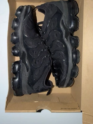 Nike Air Vapormax Plus svarta sneakers - Säljer ett par helsvarta Nike Air Vapormax Plus sneakers med ikonisk bubblig sula och mesh- samt syntetöverdel. Skorna har en sportig look med diskreta detaljer och klassisk Nike-logga på sidan. Perfekta för dig som gillar streetwear och vill ha något stilrent.