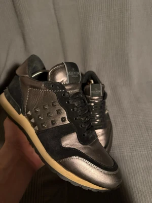 Valentino  - Snygga sneakers från Valentino i metallicgrått och svart med coola nitar på sidorna. Skorna har en mix av läder och mocka, svarta skosnören och en kontrasterande beige sula. Perfekta för dig som vill sticka ut med en exklusiv och trendig look.