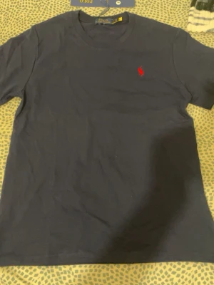 Mörkblå t-shirt från Polo Ralph Lauren - Klassisk mörkblå t-shirt från Polo Ralph Lauren med röd broderad logga på bröstet. T-shirten har rund halsringning och korta ärmar. Tillverkad i mjuk bomull som känns skön mot huden. Perfekt för en clean och stilren look.