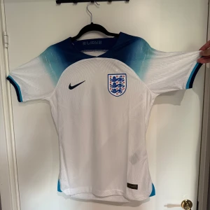 England fotbollströja - Säljer en officiell England fotbollströja från Nike med korta ärmar. Tröjan är vit med marinblå och ljusblå detaljer vid axlar och krage, samt England-emblem på bröstet. Tillverkad i lätt Dri-FIT material som andas, perfekt för match eller träning.