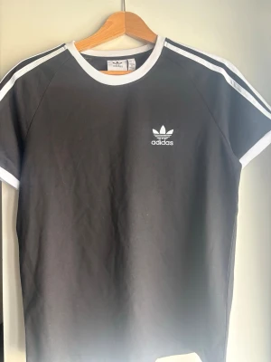 Svart Adidas t-shirt med vita detaljer - Snygg svart t-shirt från Adidas med klassiska vita ränder på axlarna och vit logga på bröstet. T-shirten har vit ribbad krage och är tillverkad i mjuk bomull. Perfekt för en sportig och avslappnad stil.