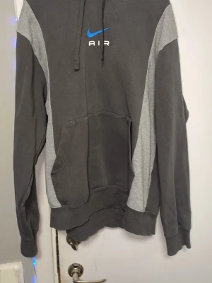 Nike Air grå hoodie med huva - Nike Air hoodie i mörkgrått med ljusgrå paneler på ärmarna och sidorna. Stor känguruficka framtill, justerbar huva med dragsko och blå Nike-logga med texten 'AIR' på bröstet. Tillverkad i mjukt bomullsmaterial, perfekt för en avslappnad streetwear-look.. står M men passar mer som L