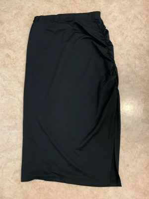 Svart långkjol med slits  - Stilren svart långkjol med rynkade detaljer på sidan och en diskret slits för extra edge. Kjolen har en enkel, rak passform och är tillverkad i ett mjukt, stretchigt material som känns bekvämt hela dagen.