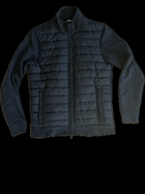 Dressman cardigan - Snygg mörkblp cardiganjacka med quiltat mönster framtill och släta ärmar. Jackan har dragkedja hela vägen, ståkrage och två fickor med dragkedja. Perfekt för lager-på-lager och en clean look.