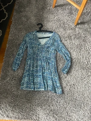 Blå mönstrad klänning från Zara - Supersnygg blå kortklänning från Zara med vitt mönster och v-ringning. Klänningen har långa ärmar och en flowy passform som ger en avslappnad vibe. Perfekt för varmare dagar och enkel att styla med sneakers eller boots.
