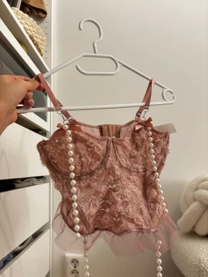 Rosa spets bodyset med pärldetaljer - Sött tvådelat underklädesset i ljusrosa spets med 3D-blommor, volangkant och dekorativa pärlband. Axelbanden är justerbara och både topp och trosa har små rosetter. Materialet är genomskinligt mesh och spets, vilket ger en romantisk och lekfull look. Aldrig använt enbart tvättat. 