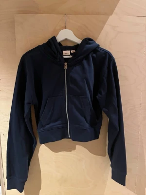 Mörkblå hoodie med dragkedja - Oanvänd mörkblå hoodie i mjukt bomullstyg med dragkedja framtill. Modellen har huva och ribbade muddar vid ärmslut och nederkant. Perfekt för en avslappnad och stilren look. 