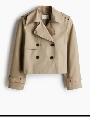 Kort beige trenchcoat med dubbelknäppning - Stilren kort trenchcoat i beige med dubbelknäppning och bred krage. Jackan har axelklaffar, dekorativa knappar och justerbara spännen vid ärmsluten. Perfekt för en trendig och klassisk look. Materialet ser ut att vara bomull eller bomullsblandning.